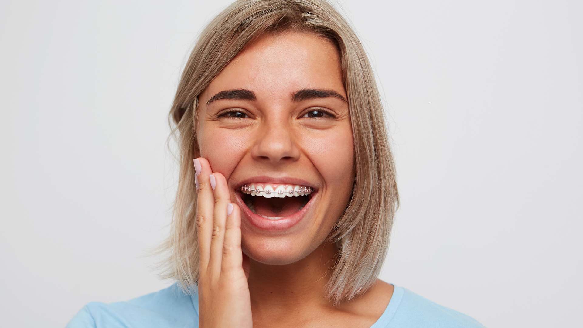 Cheerful beautiful young woman with blonde hair and braces on teeth laughing and touching her face by hand isolated over white background
